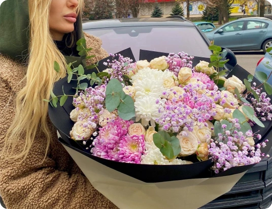 Жінка тримає великий букет з білих, рожевих і бузкових квітів на тлі припаркованих автомобілів